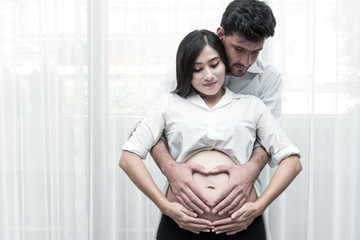 Fototapeta premium Mixed race parenthood concept. Portrait of young Chinese pregnant and her white male husband holding each other. Taken indoor with real six months old pregnant woman.