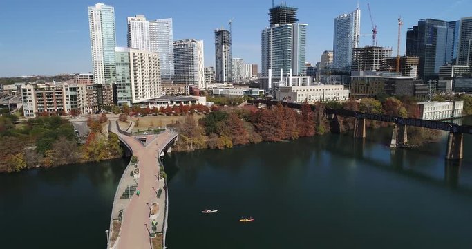 A slow rising aerial establishing shot (DX) of the Austin city skyline with the Pfluger Pedestrian Bridge and kayakers in the Colorado River in the foreground on a late sunny Autumn day.  	
