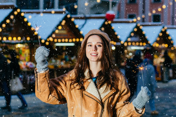 Amazing woman hat walking on the street with sparkler. Adorable female woman in winter outfit spending time outdoor and looking at Bengal light over Festive Christmas fair on background.