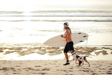 Surfer in reef boots with surfboard and helmet running jogging on the beach with his dog at sunset in Bali, Indonesia.