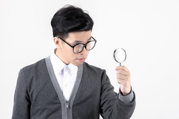 Nerd studying hard. Young Asian nerd man wearing glasses holding magnify glass standing isolated on white background. Black hair, grey sweater, white shirt. Education concept.