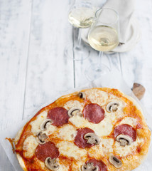 pizza with salami and mushrooms on a white wooden background and two glasses of white wine