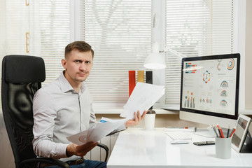 Shocked perplexed and stress business man in shirt sitting at the desk, working at computer with modern monitor, folders, coffee, documents in light office on window background. Manager or worker.