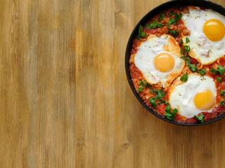 A pan of fried eggs with tomato sauce and parsley on a wooden background. Shakshuka a traditional meal of the Jewish cuisine