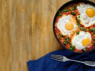 A pan of fried eggs with tomato sauce and parsley on a wooden background. Shakshuka a traditional meal of the Jewish cuisine
