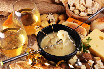 Gourmet Swiss fondue dinner on a winter evening with assorted cheeses on a board alongside a heated pot of cheese fondue with two forks dipping bread and white wine behind in a tavern or restaurant