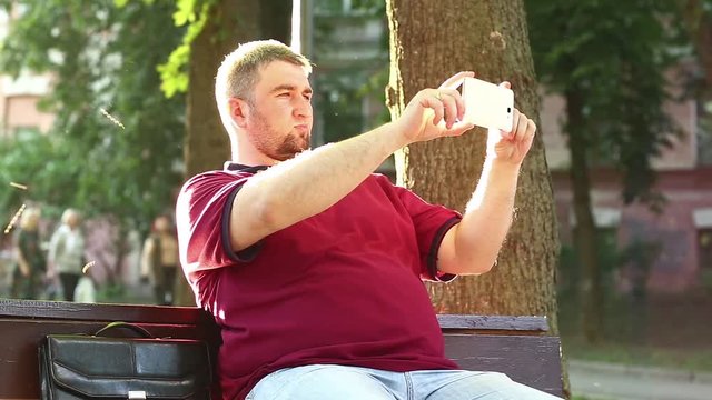 Businessman sits on the bench and records videos on smartphone. Fat man makes photos on his cell phone