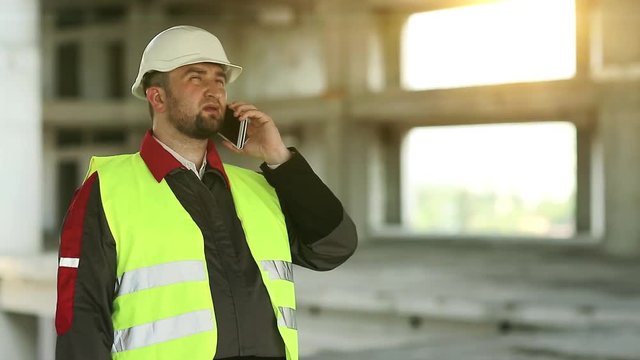 Builder talks on cell phone on construction site. Workman in green vest and white hard hat speaks on smartphone on project site