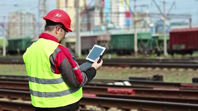 Railwayman with computer at freight train terminal. Railway employee makes notes in his tablet pc. Railwayman in uniform and red hard hat works with tablet computer