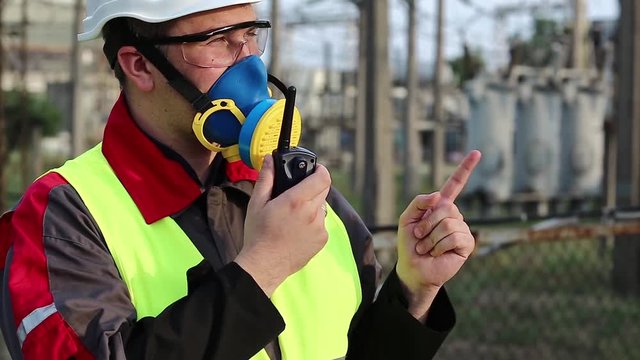 Powerman in respirator and with portable radio transmitter on electric power station. Power engineering specialist in gas mask, goggles and hard hat on heat power plant communicates via radio station