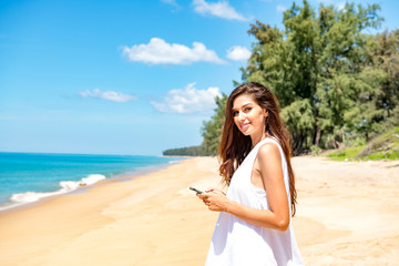 Fototapeta na wymiar Beautiful young single white woman on beach. Using mobile phone calling her friends, wearing white dress. Connected world concept.