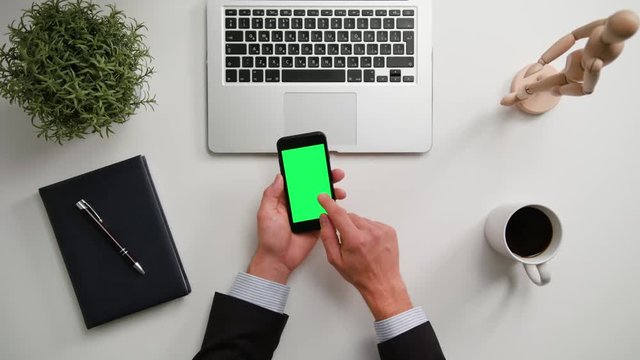 A man's hands holding a phone and touching the green sreen over a white table. View from the top. Close-up shot