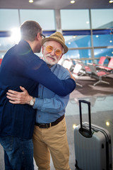 Sweet bonding. Pleasant elderly bearded father and his adult son are standing at international airport. They are hugging while feeling happiness. Back view of guy