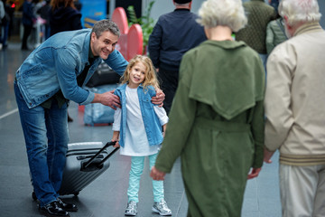 Fototapeta premium Say goodbye. Grandparents are accompanying adult son and his daughter to airport. They are standing back to camera. Focus on positive man hugging little girl with suitcase