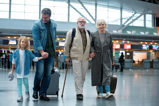Full length of delighted old grandparents are going with their son and granddaughter at airport terminal to check-in on their flight. They are carrying suitcases and expressing happiness