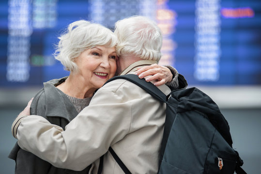 Happy to see you. Positive charming romantic aged couple are standing against electronic board at international airport and hugging while feeling happiness. Back view of man with backpack