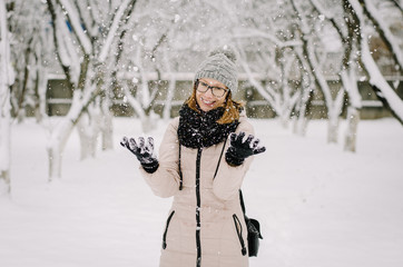 Young attractive girl in nerd glasses, a beige down jacket and knitted hat with a pompon smiles and fools around, throwing snow with her arms. Closeup shot.