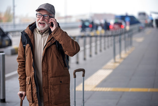 Deeply involved in conversation. Portrait of joyful senior man with beard is resting on walking stick and talking on smartphone with smile. He is standing outdoors with his suitcase. Copy space