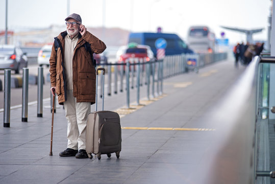 Glad to hear you. Full length portrait of cheerful bearded gray-haired man in glasses is resting on cane while standing outdoors and talking on mobile phone. He is expressing gladness. Copy space - Powered by Adobe