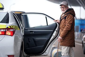 Portrait of positive bearded old man in glasses is going to sit into the car. He is opening door of...