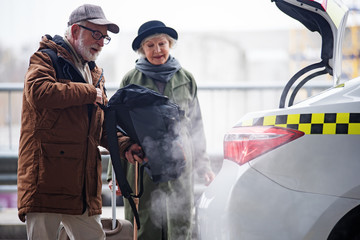 Pleasant gray-haired man in glasses is putting his backpack in trunk of taxi while standing together with his aged wife. Focus on male. Copy space in the right side