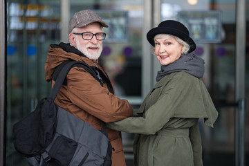 Feeling happy. Portrait of optimistic stylish aged couple is standing together by hand. They are looking at camera with joy while going to the airport building