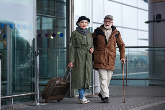 Arrival. Full length portrait of pleasant positive senior man with cane and woman with suitcase are standing together near airport building. They are looking at camera with slight smile