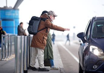 Need transport. Full length side view of senior couple is standing near road. Profile of positive bearded old man in glasses and with backpack is raising hand while stopping car