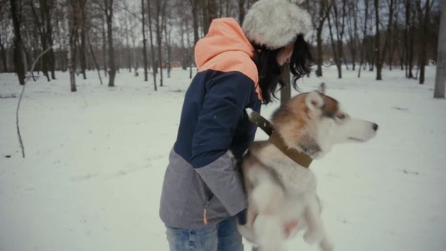 Young woman is holding a hussel dog in her arms in a snowy winter park. Beautiful girl playing with a dog