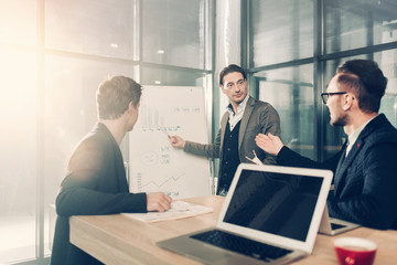 Serene man demonstrating scheme to young colleagues while standing near it. They sitting at table in office. Presentation concept