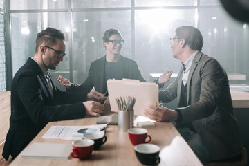 Outgoing man flourishing arm while speaking with colleagues. They sitting at table. Serene adult watching at laptop. Business concept