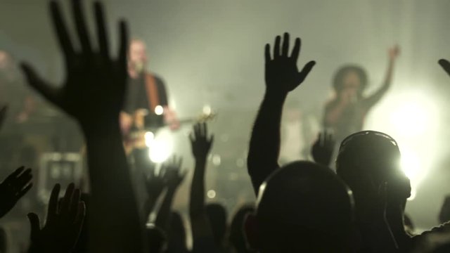 Stock video footage Crowd Raising hands at a concert, focus is on the hands, band is out of focus in background. silhouetted people. 