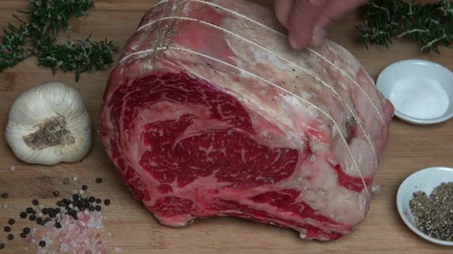 Raw Aged Marbled Prime Rib on a Wood Cutting Board Being Seasoned with Salt and Pepper