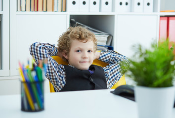 Small caucasian curly boy looks at the camera waved his hands behind his head and leaned back in his chair. Boy playing in the boss at the office.