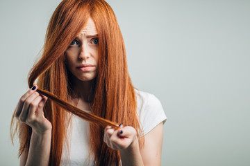 Sad redhead girl holding her damaged hair looking at camera