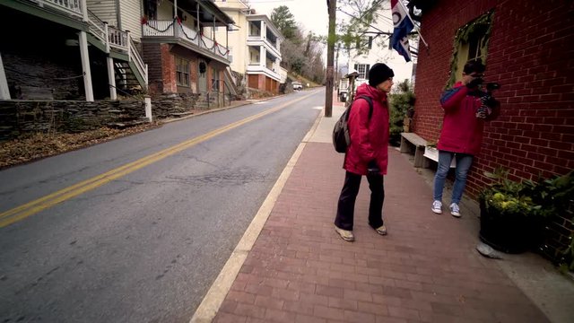 Mature woman walks down a sidewalk and waits for her teenage vlogging son to photograph in Harpers Ferry, West Virginia.