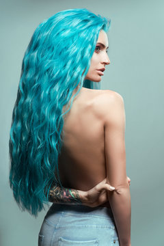 Beautiful woman with curly blue hair and tattoos posing at camera against blue background, back view