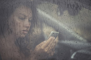 Sad woman looking in the mobile phone and reading message in the window with rain drop in the car. Face of young female behind rain car window. Loneliness and depression concept. Psychology