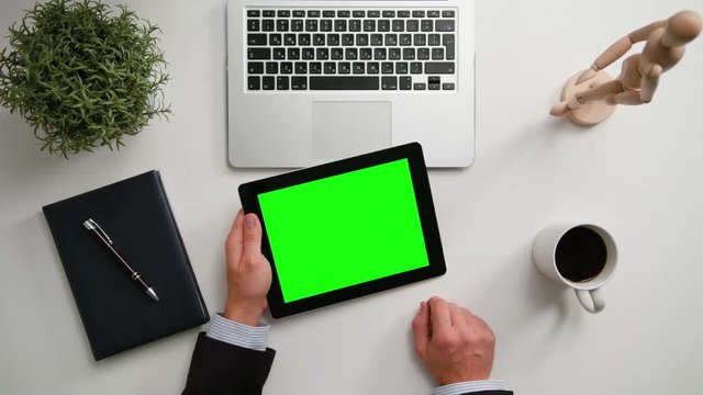 A man's hands holding an i-Pad and touching the green sreen over a white table. View from the top. Close-up shot.