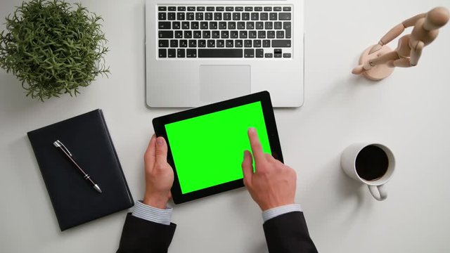 A man's hands holding an i-Pad and touching the green sreen over a white table. View from the top. Close-up shot.