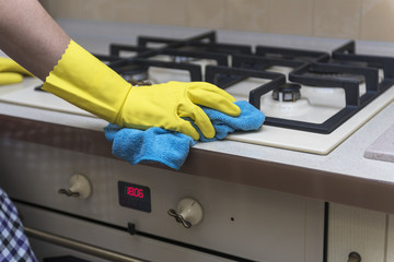 Women's hand in protective gloves wipes the kitchen gas stove with a blue rag.