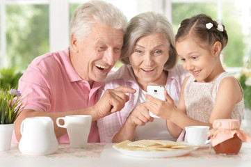 Obraz na płótnie Canvas Smiling grandparents with granddaughter using smartphone