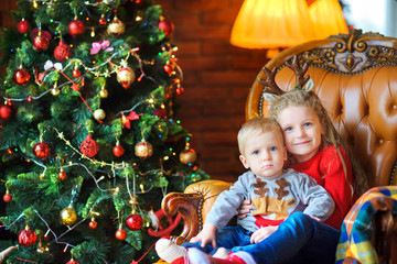 sister gently hugs her little brother sitting in a chair near a festive Christmas tree.