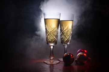 New Year Eve celebration background with pair of flutes and bottle of champagne with Christmas attributes (or elements) on snowy dark toned foggy background. Selective focus.