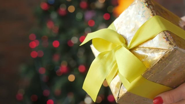 Close up Gift, female hands hold and show the gift, the golden box is bandaged with yellow tape. Bright festive background