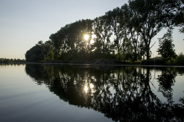 Trees on the shore are reflected on the surface. The sun is making its way to the lake through the branches.  Cod and dry branches the right border the coast and water. New life is on the horizon.