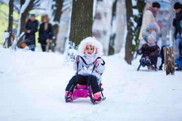 Fototapeta na wymiar Pretty smiling little girl in her ski suit sliding down a small snow covered hill with her sledge board during a bright sunny winter day