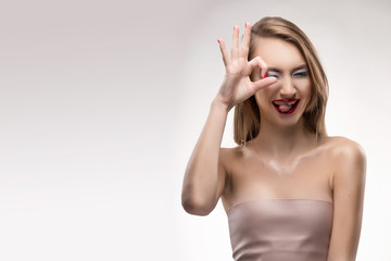 Fototapeta premium The beautiful blonde red lips smiling girl shows the sign ok, squints and flicks out tongue.