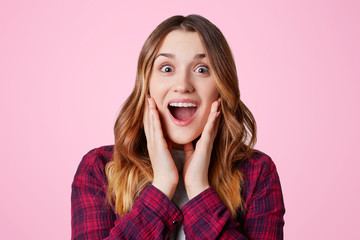 Photo of excited pretty woman looks in amazement, bugged eyes and widely opened eyes, being pleasantly shocked to recieve present, isolated over pink backgroud. Emotional female poses indoor
