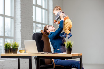 Young multitasking businessmam dressed in the suit having fun with her baby son at the office
